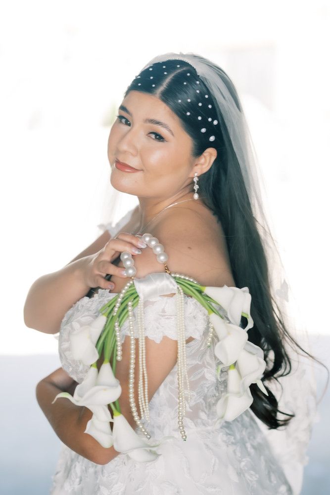Bride wearing pearl-studded headpiece and white gown holds calla lilies in romantic portrait.