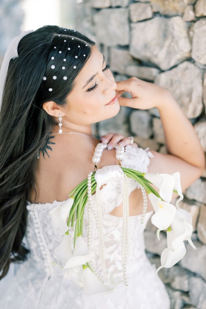 Elegant bride in white dress with pearl headpiece poses against textured stone backdrop in artistic portrait.