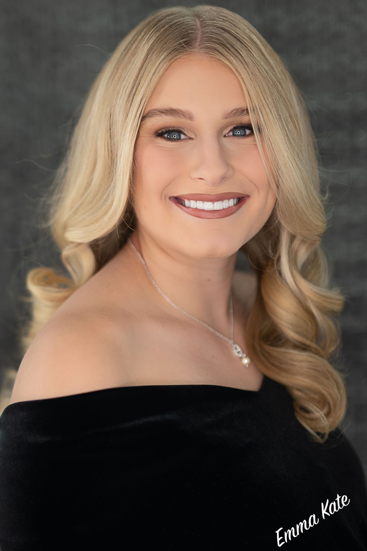 Young blonde woman in black off-shoulder top smiles warmly, wearing a delicate necklace in a professional portrait photo.