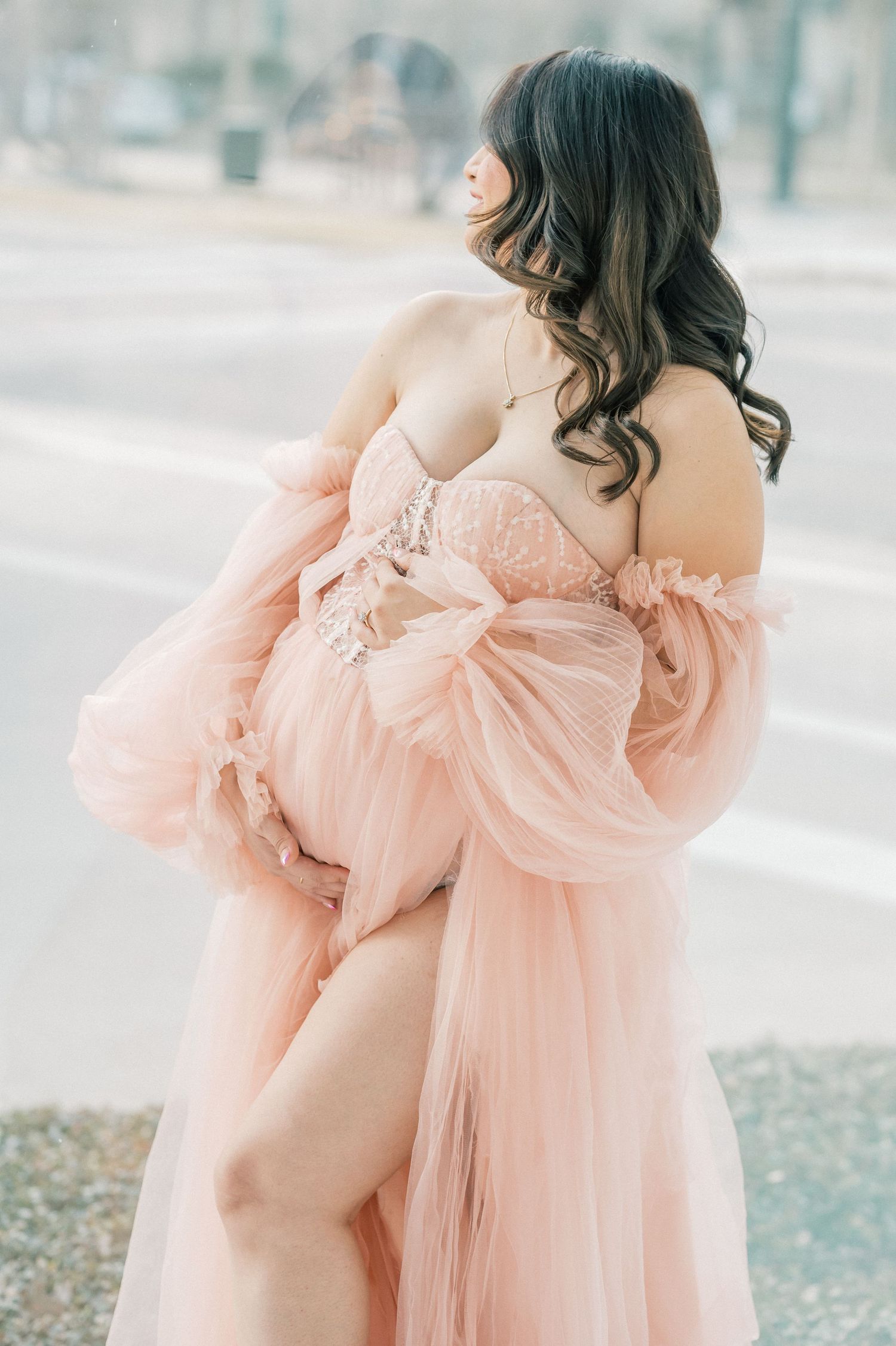 Pregnant woman in blush pink tulle robe and lace corset gown cradling baby bump outdoors.