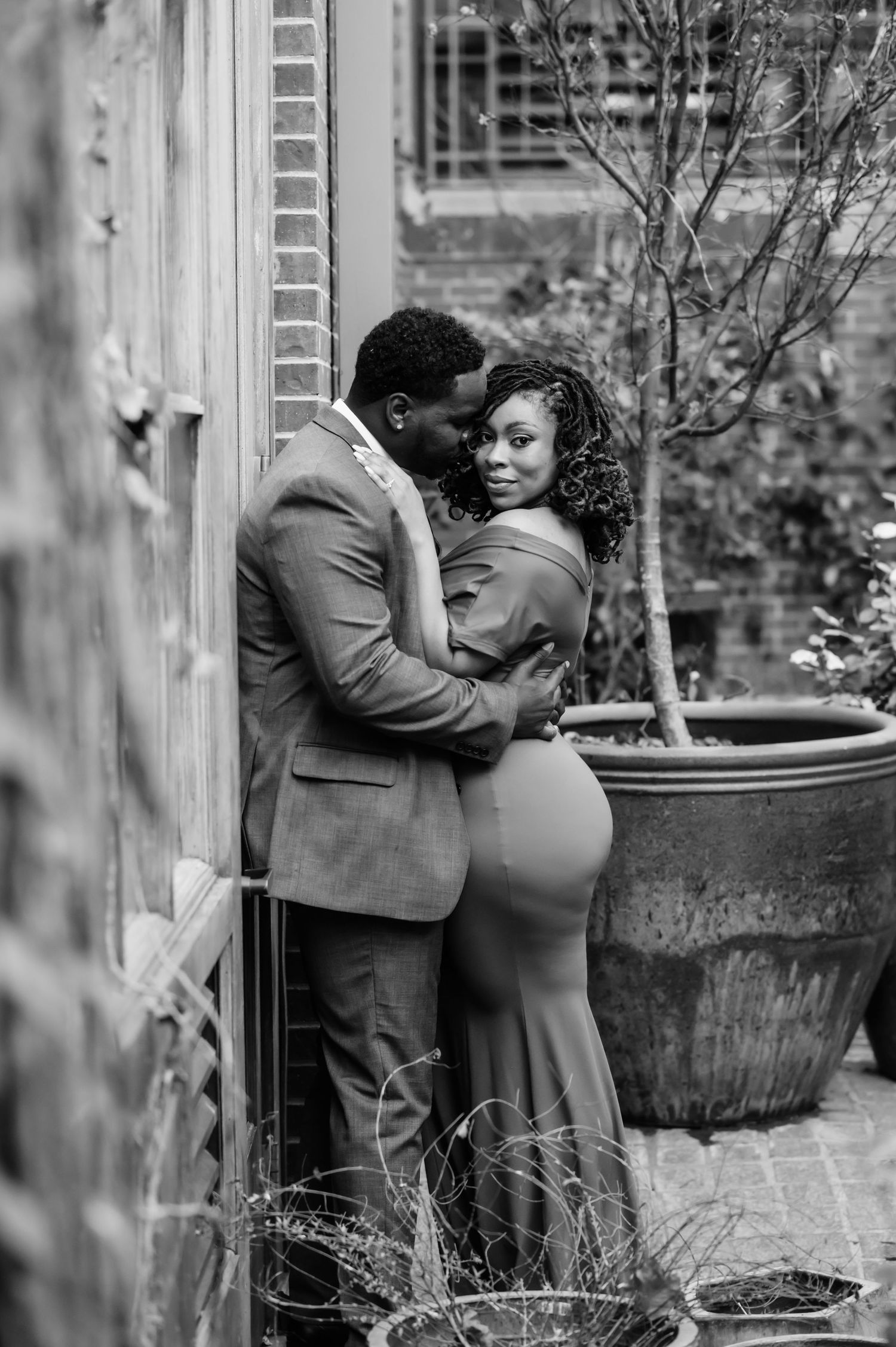 Black and white photo of an elegant couple embracing romantically against a brick wall in a lush garden setting.