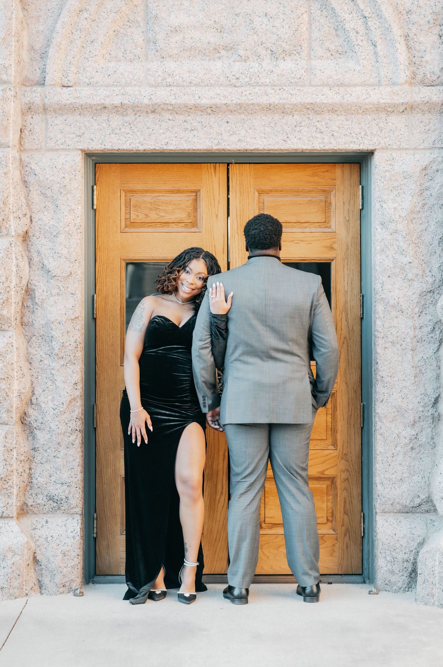 Couple posing at ornate wooden doors, woman in black velvet slit gown leaning against man in gray suit.