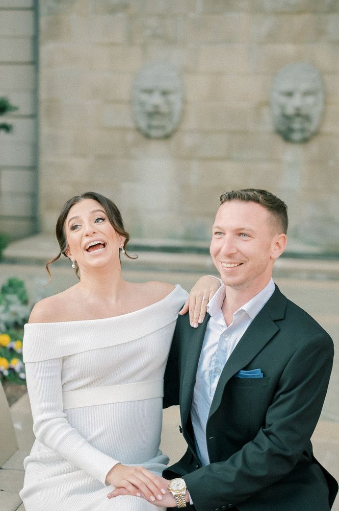 Laughing bride in off-shoulder white dress and groom in dark suit sitting together before stone facade.