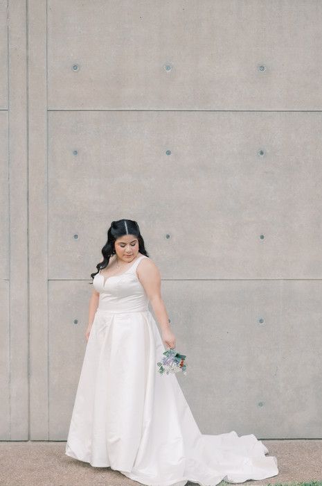 Bride in white A-line gown with train holds bouquet against concrete wall.