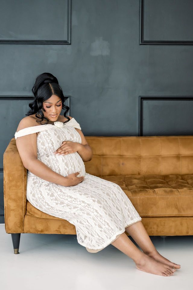 Pregnant woman in white lace dress cradling baby bump while seated on tan velvet sofa against dark panel wall.