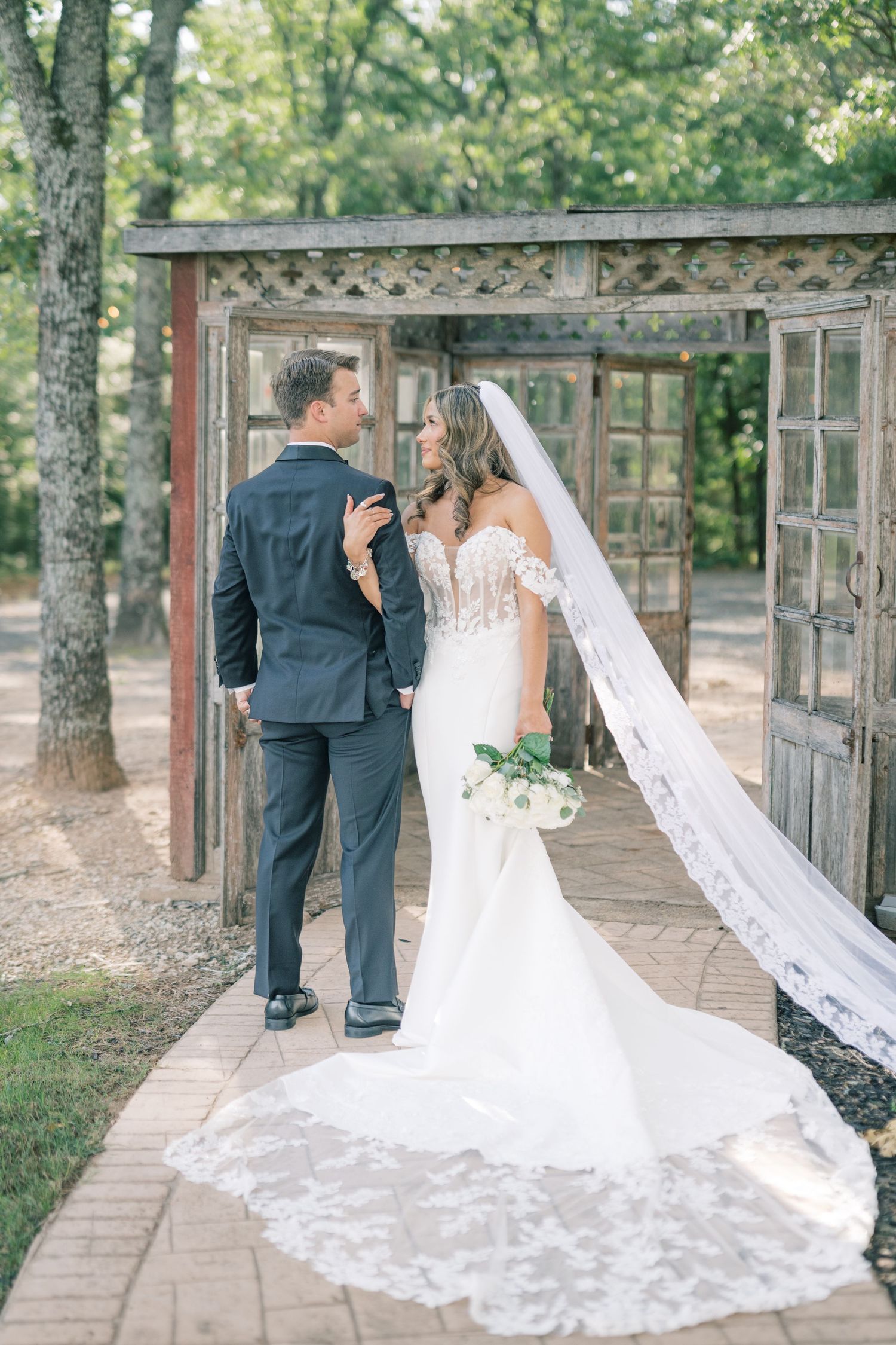 Bride and groom sharing romantic moment under rustic wooden arbor surrounded by lush green trees outdoors.