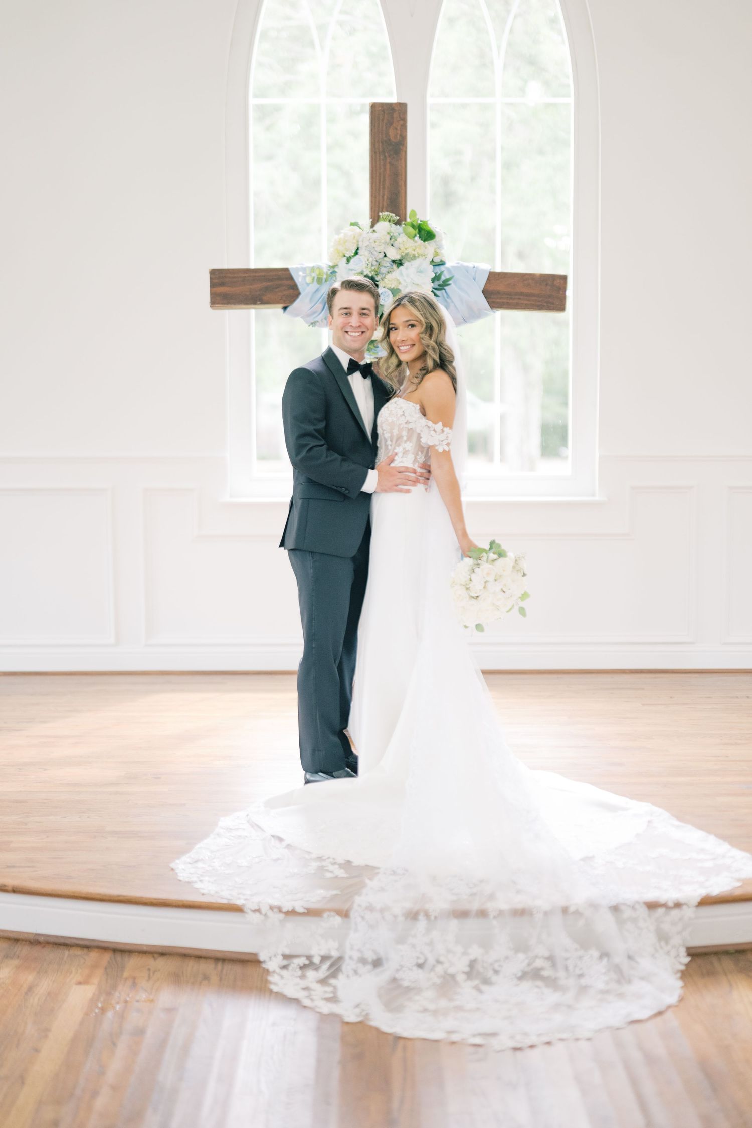 Bride and groom posing before wooden cross with white floral decor in bright chapel after ceremony.