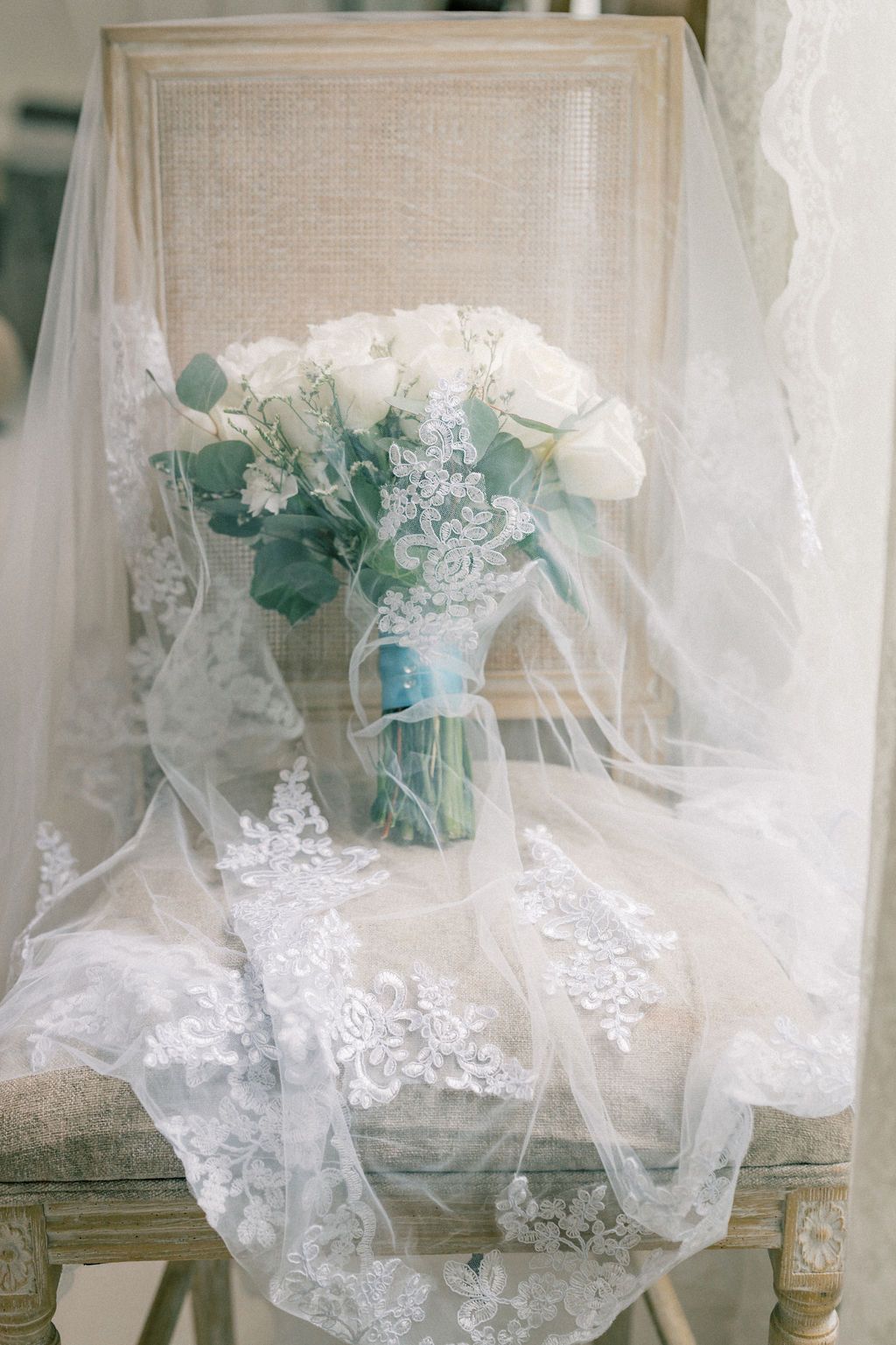 Elegant bridal bouquet of white roses in a mason jar draped with lace wedding veil on a vintage chair.