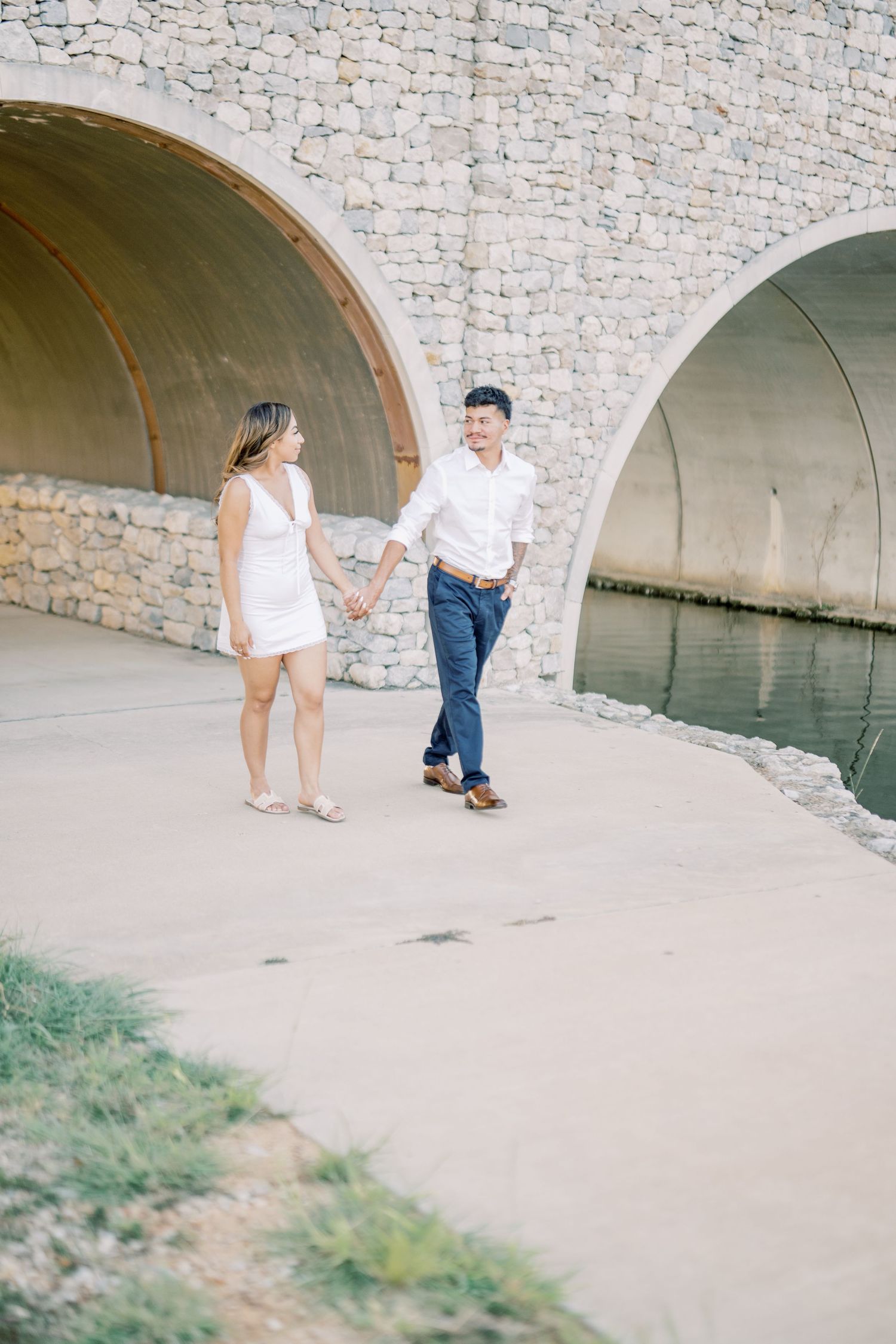 Couple walking hand in hand along waterway beneath stone bridge arches.