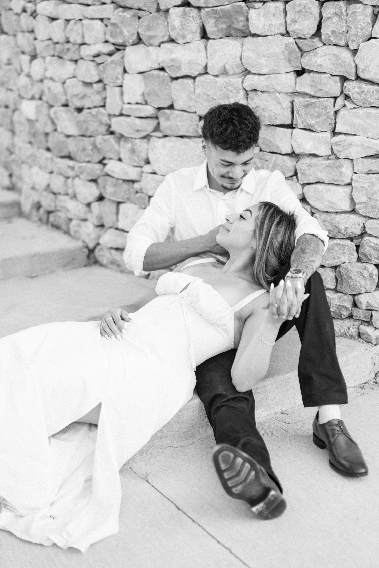 Black and white photo of couple sitting against stone wall in romantic pose.