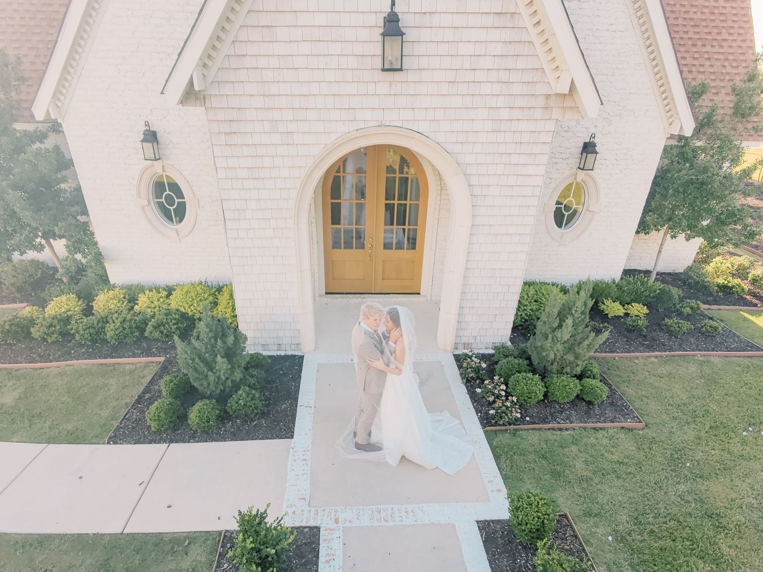 Aerial drone view of bride and groom kissing at arched chapel entrance with manicured gardens.