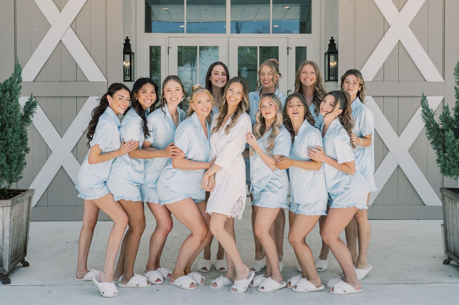 Bride in white robe poses with bridesmaids in matching light blue pajama sets outside barn venue.
