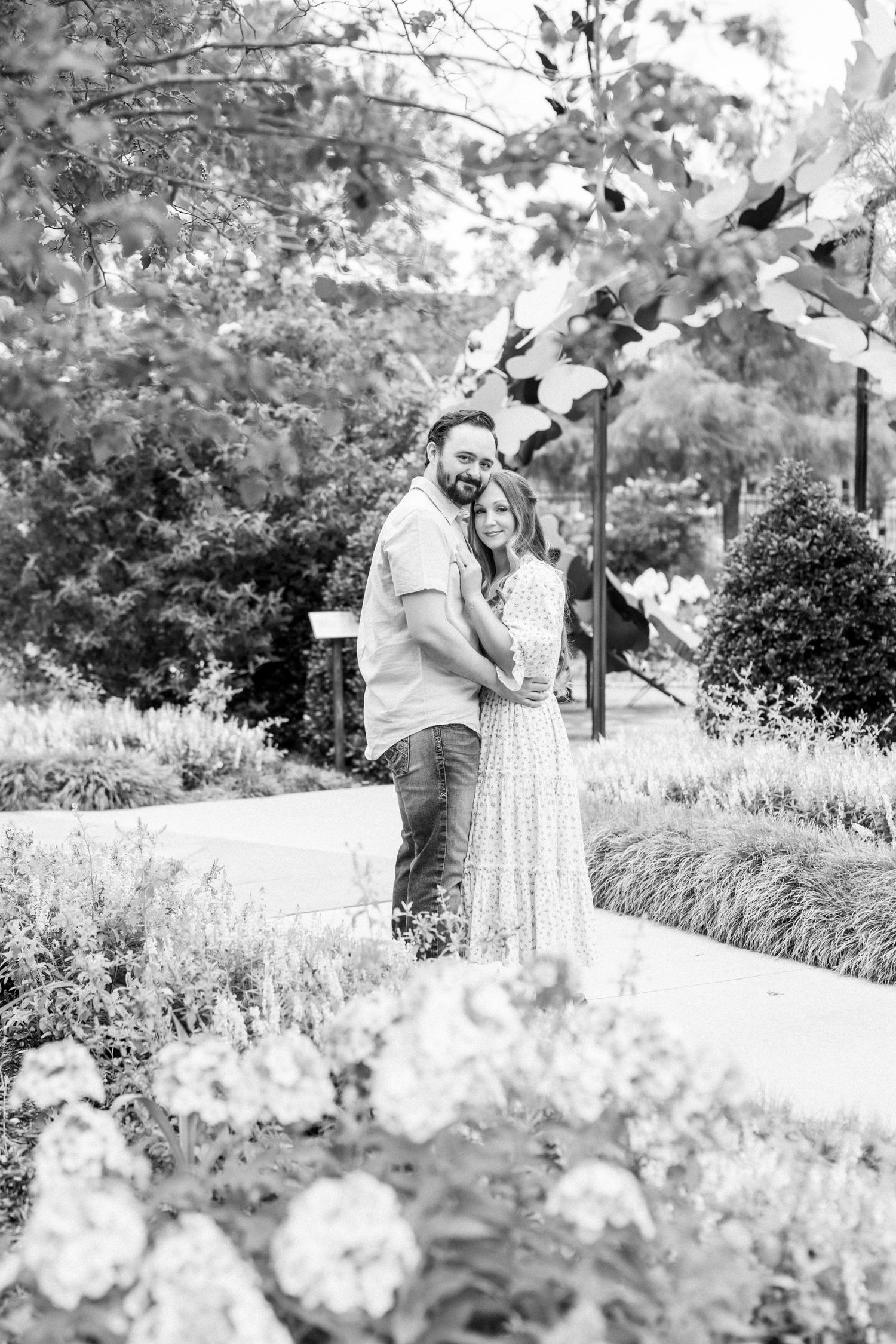 Black and white photo of a couple embracing in a lush garden with flowers in the foreground.