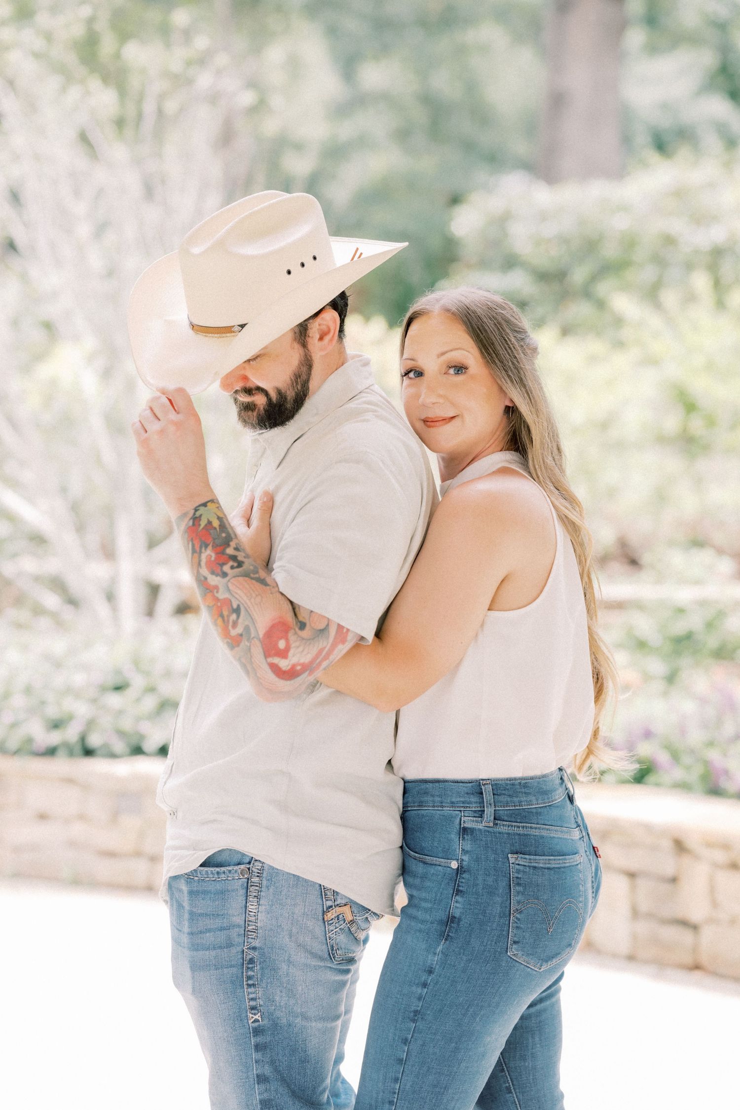 Woman leans against tattooed man wearing a white cowboy hat and jeans in a sun-dappled outdoor setting.