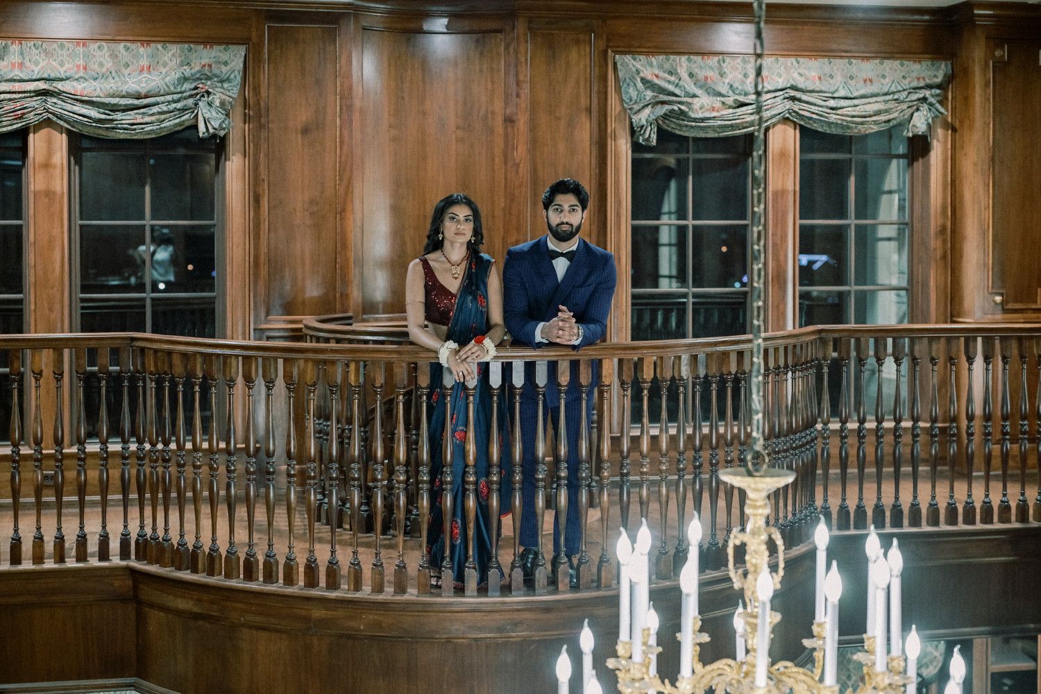 Couple poses on curved wooden balcony with chandelier in elegant indoor setting.