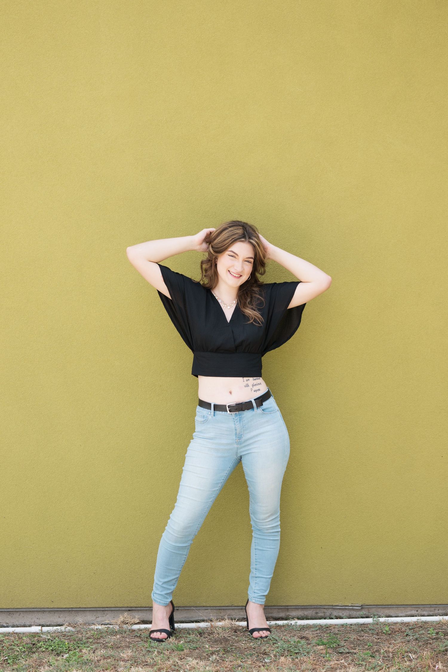 Young woman in black crop top and light blue jeans posing against a yellow-green wall with hands in hair.