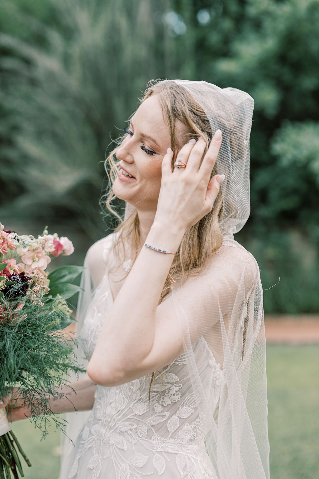 Smiling bride in lace gown and veil holding bouquet, touching hair with ringed hand outdoors.