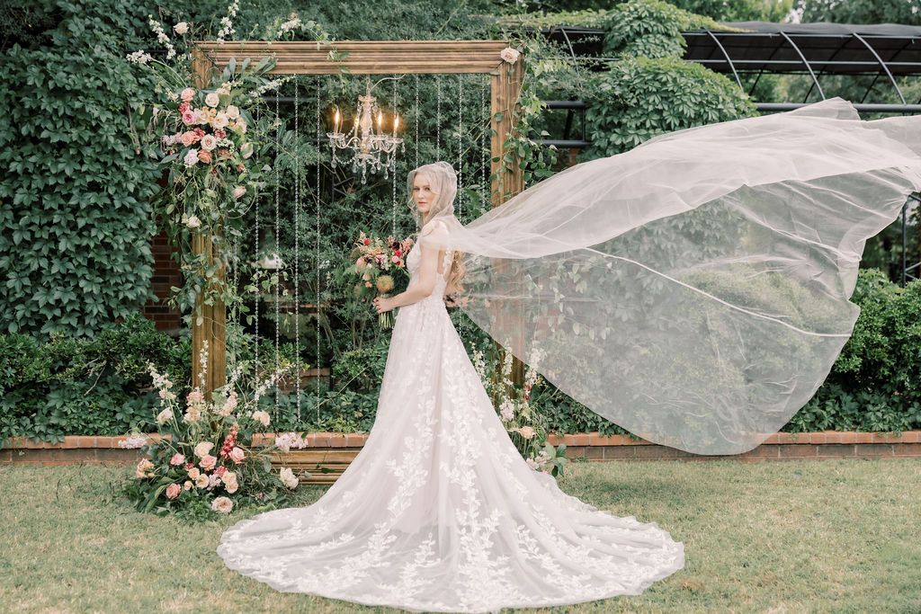 Bride in lace wedding gown with flowing veil billowing in wind, standing before floral arch with chandelier in garden.