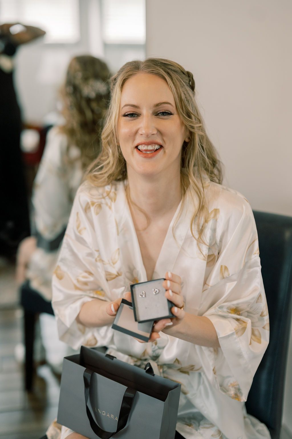 Smiling bride in floral satin robe holding jewelry box and gift bag while getting ready for wedding.