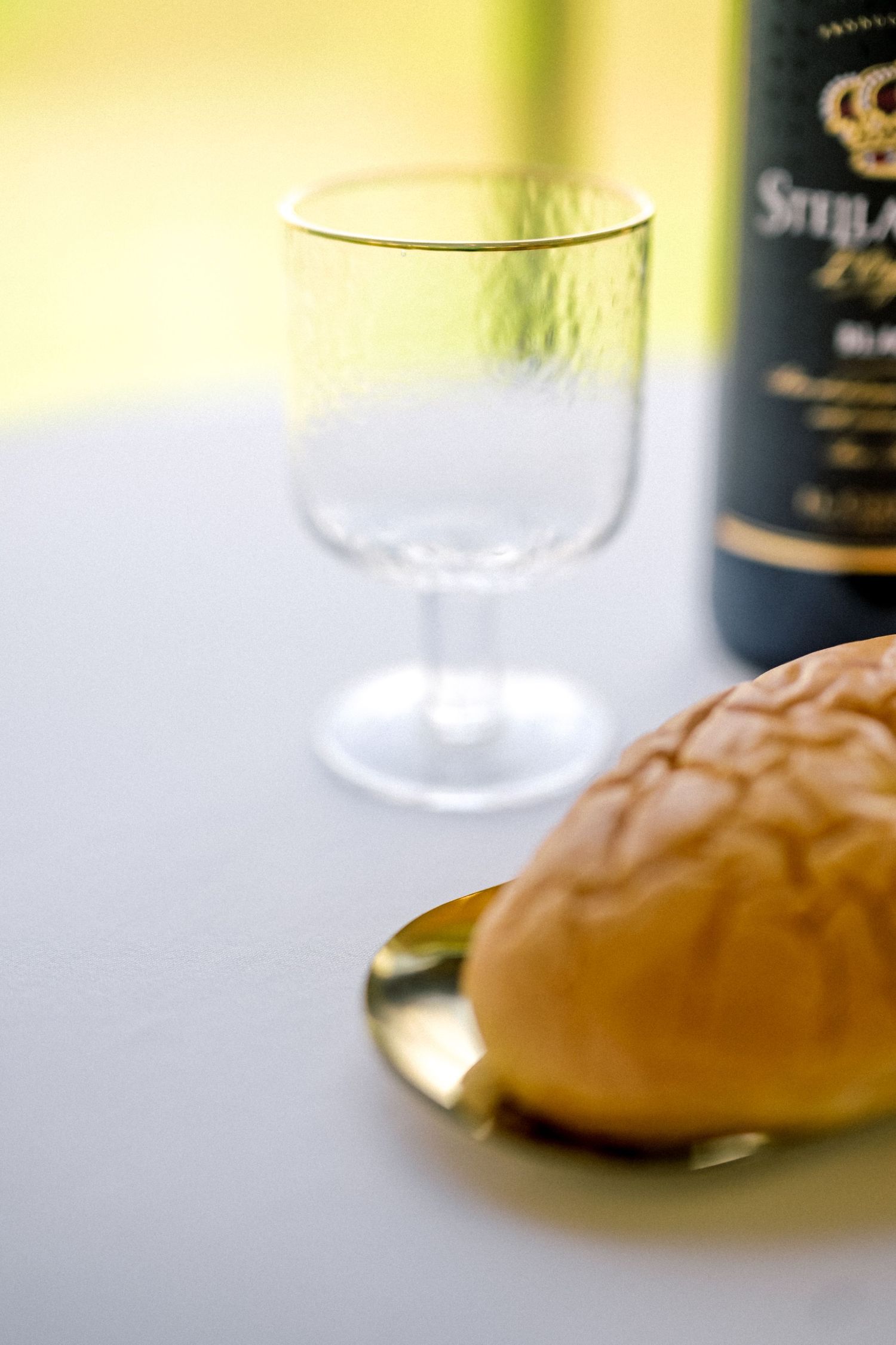 A communion bread roll on a gold plate beside an empty crystal goblet and a dark wine bottle on a white tablecloth.