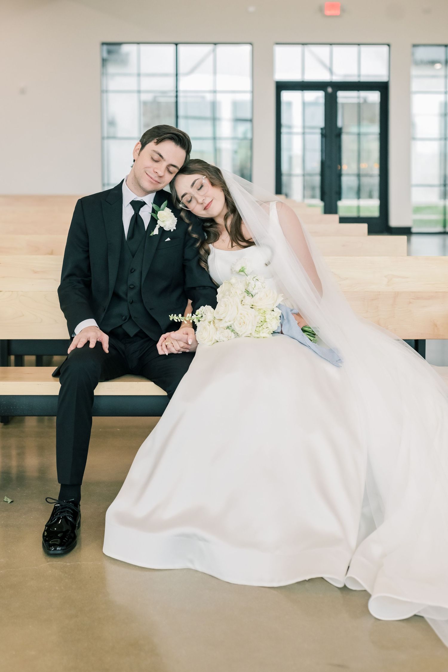 Newlywed couple in black suit and white gown resting heads together on wooden bench after ceremony.