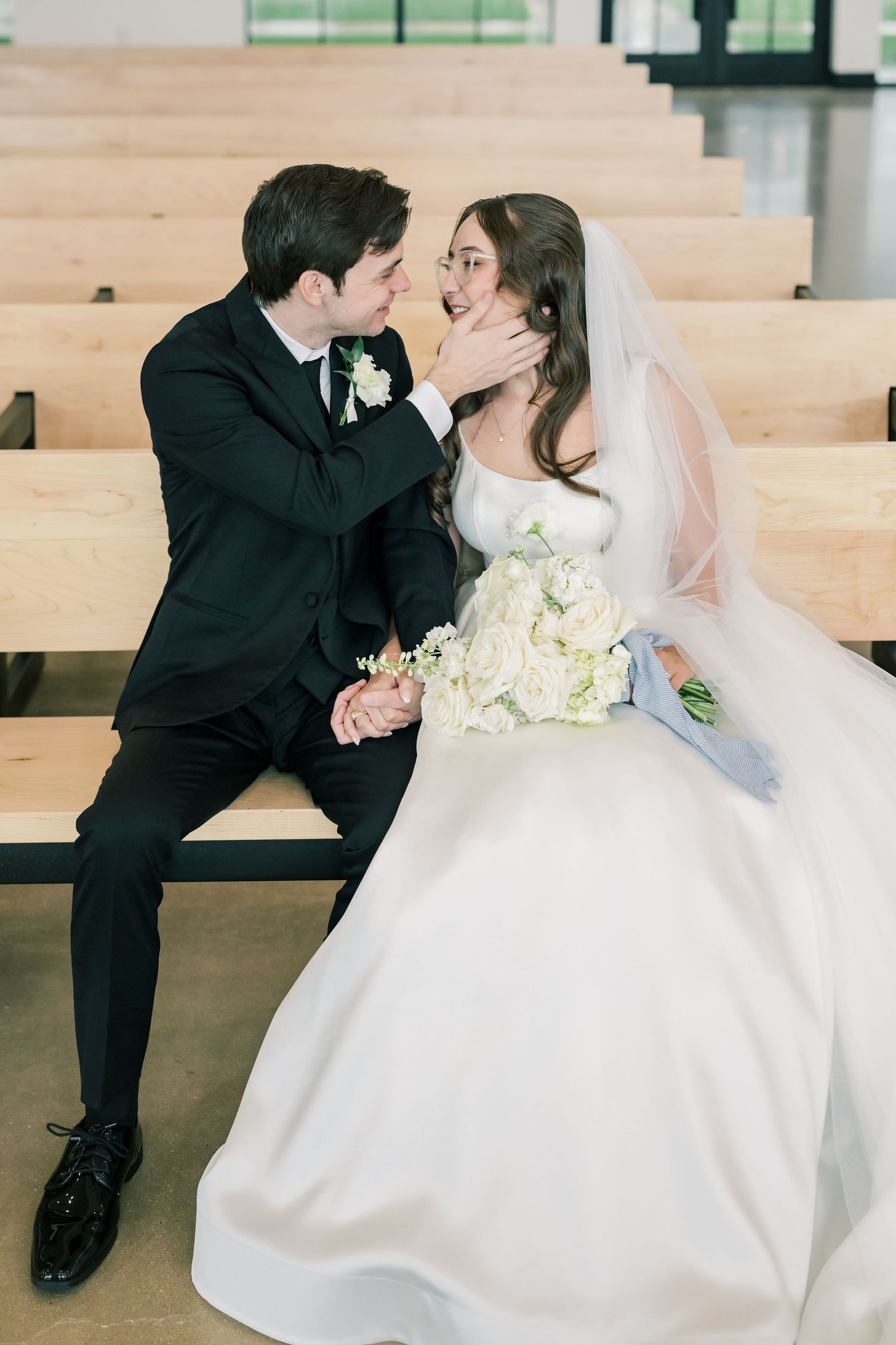 Bride and groom sharing romantic moment seated on wooden church pew, bride holding white bouquet.