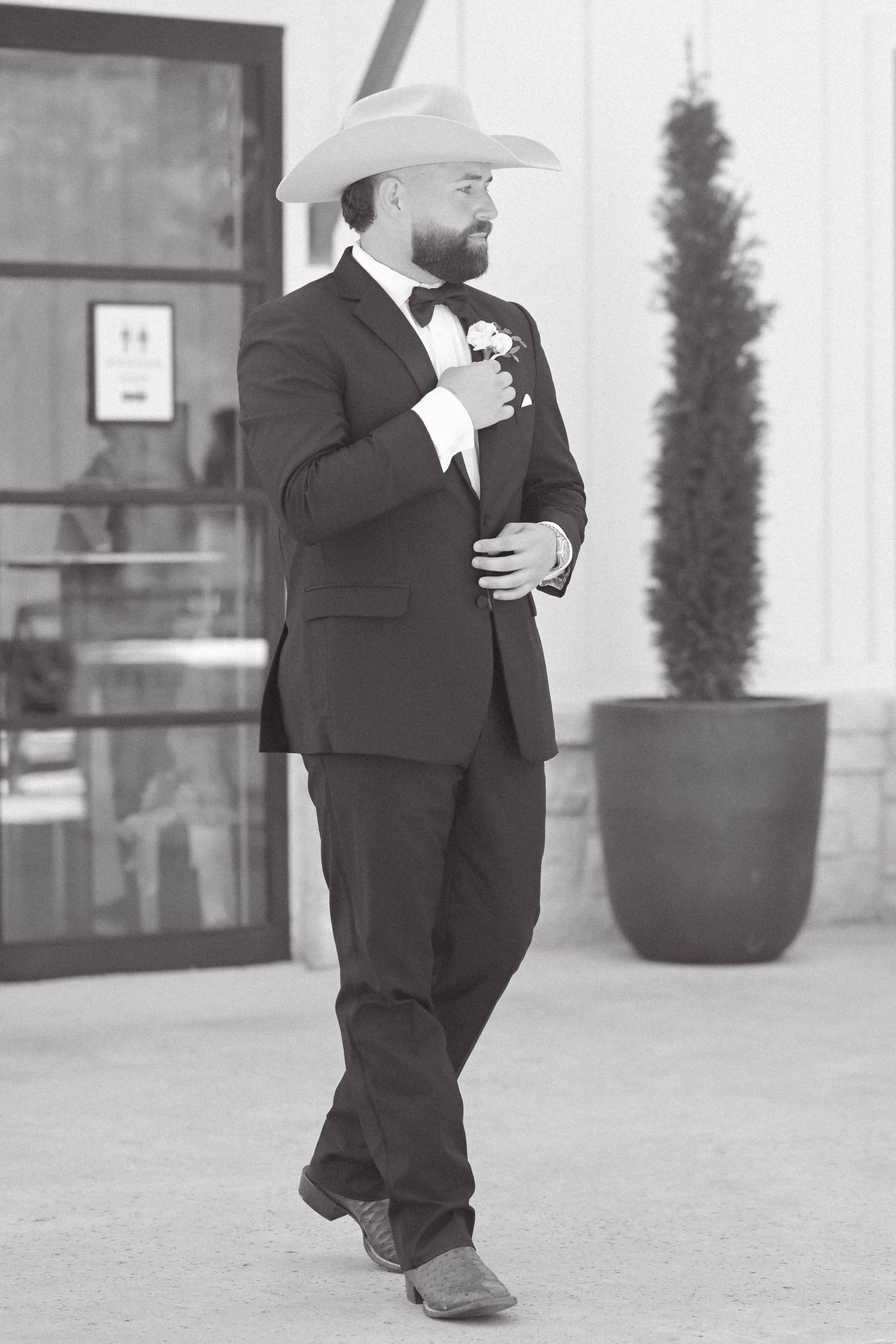 A bearded groom in a black tuxedo, cowboy hat, and boots stands confidently outside a venue.