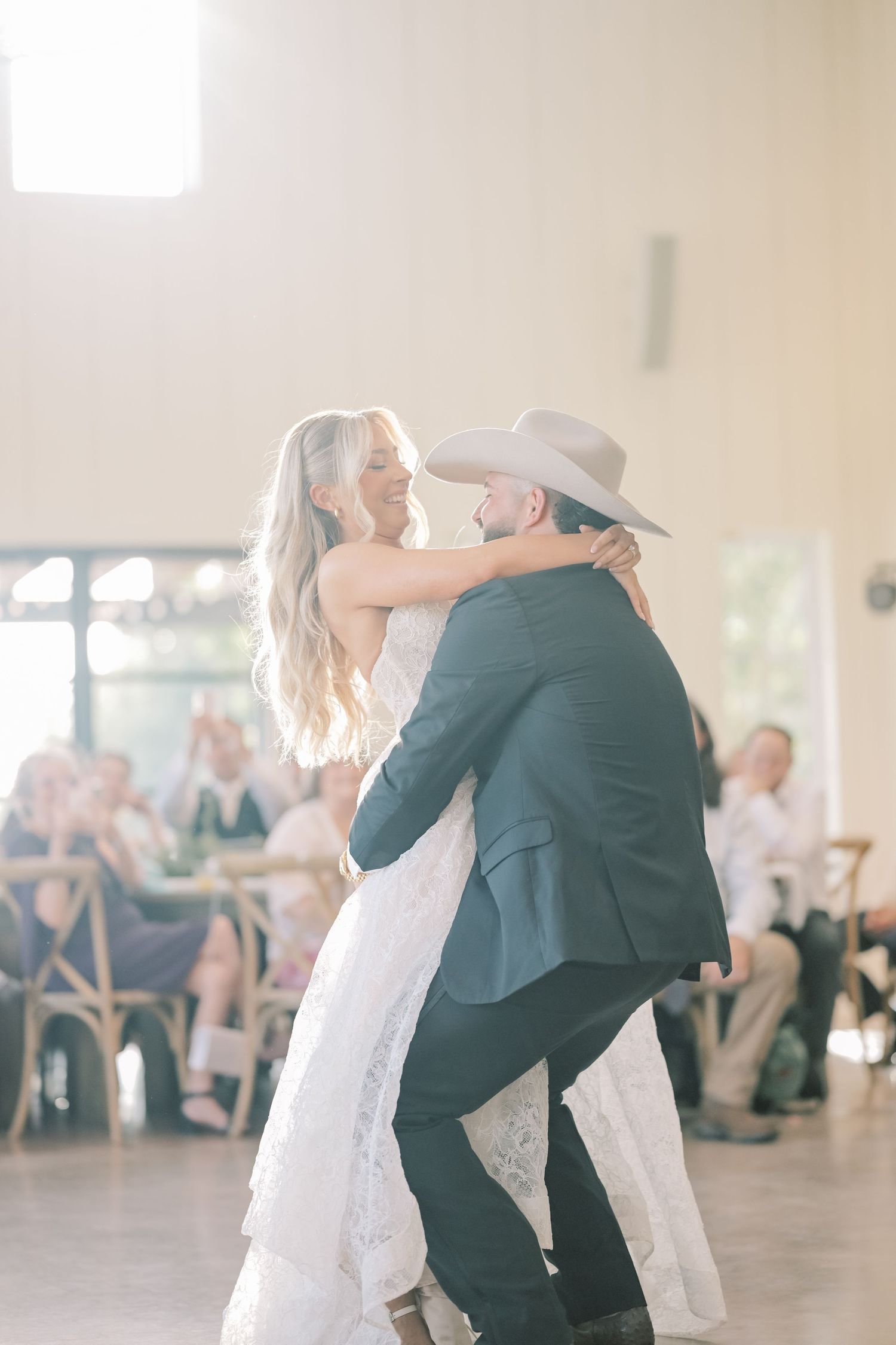Bride and groom share first dance in sunlit reception hall while guests watch in background.