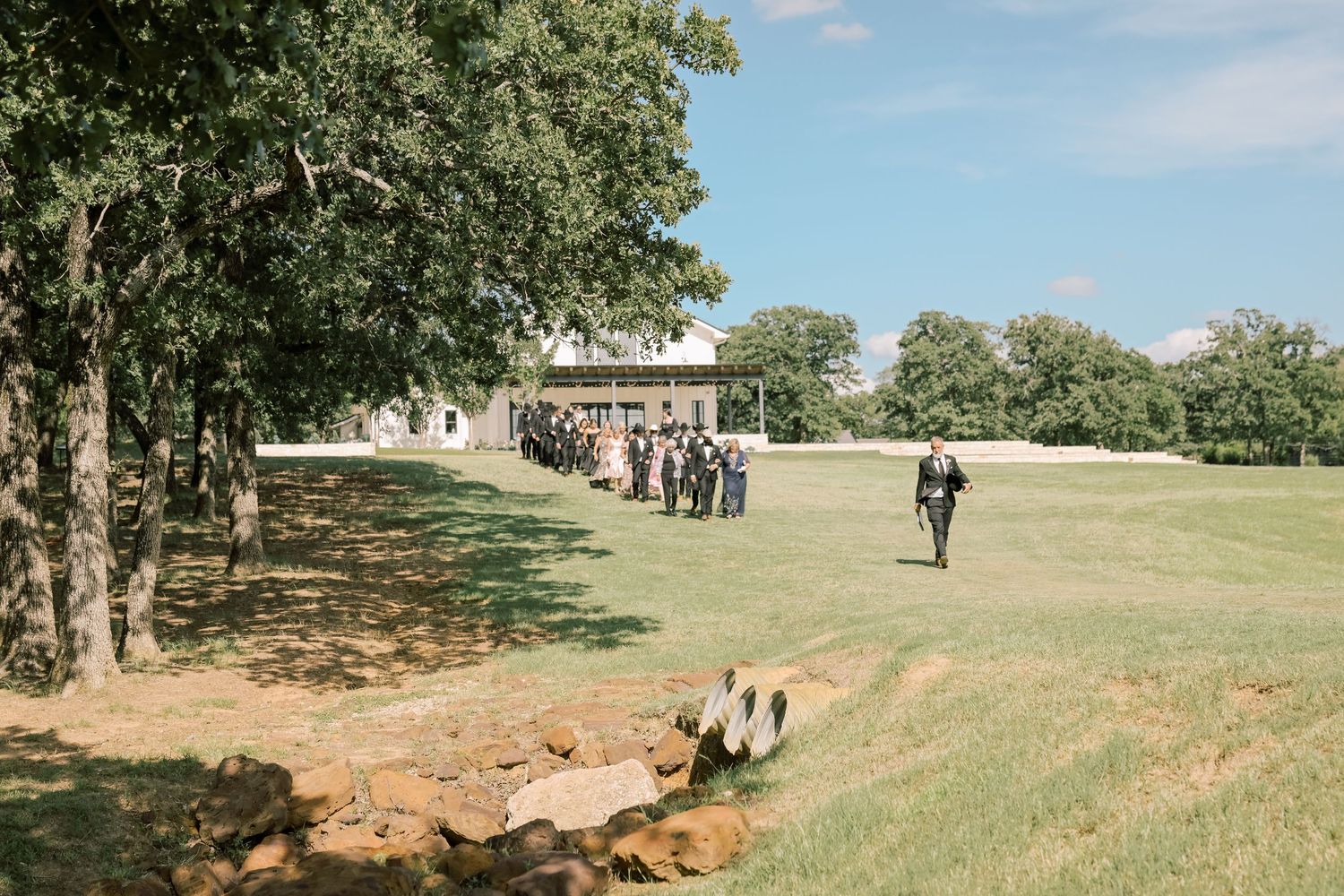 Outdoor wedding ceremony setup on green lawn with guests gathering near rustic white farmhouse venue.