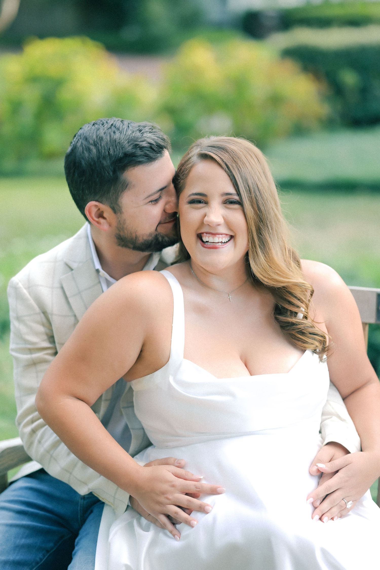 A smiling woman in a white dress laughs while a bearded man in a blazer kisses her cheek on a park bench in a green garden.