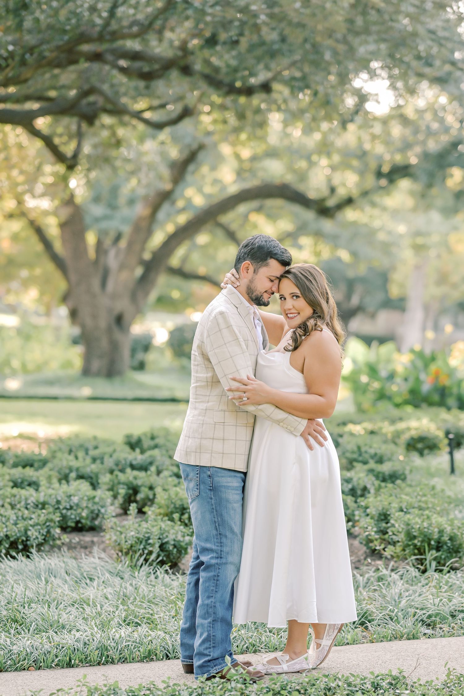 Smiling couple in a white dress and plaid blazer embrace near neatly trimmed hedges and a large oak tree at a park engagement session.