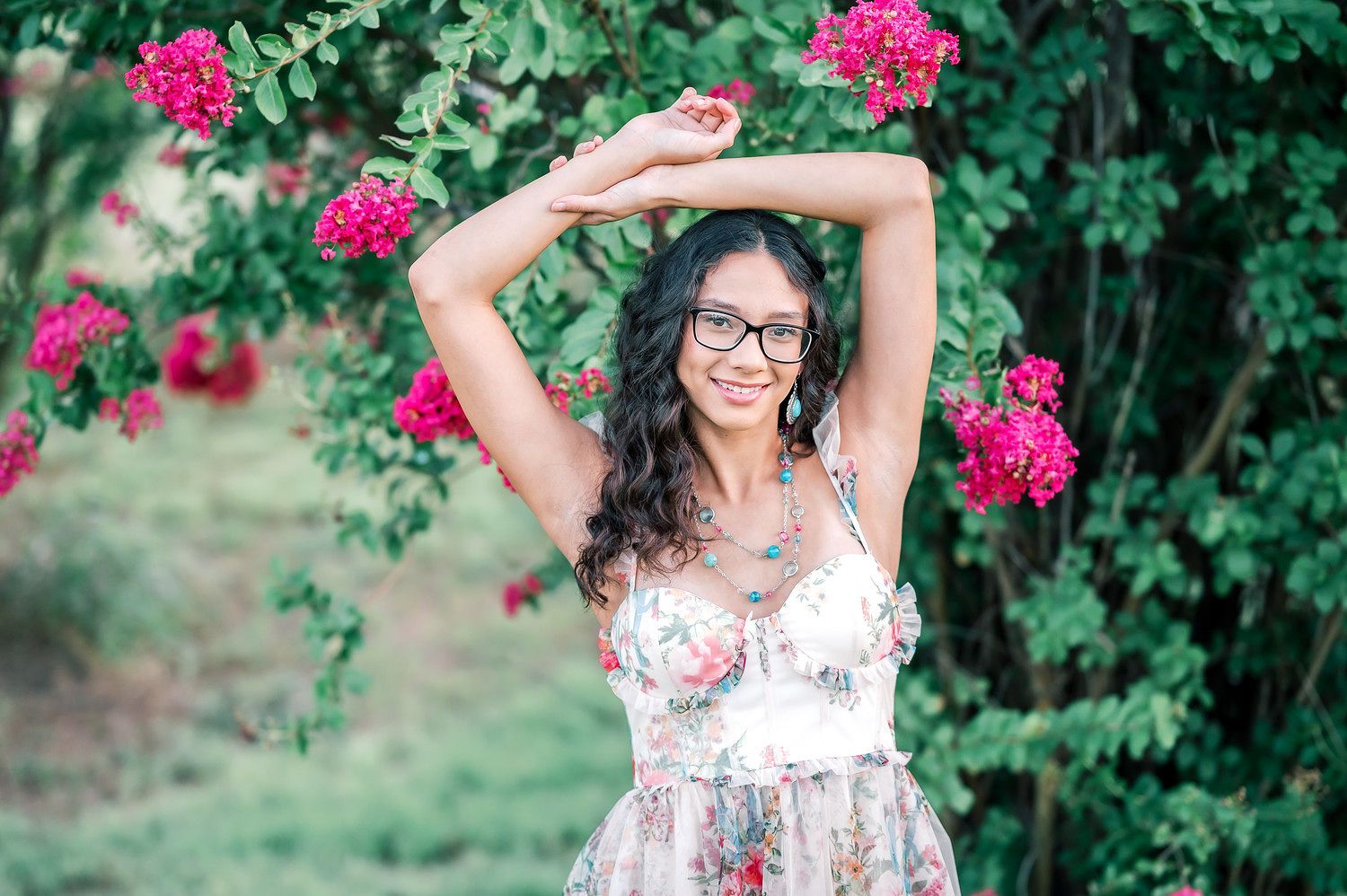 Young woman in floral dress and glasses poses gracefully under blooming pink roses in a lush garden.