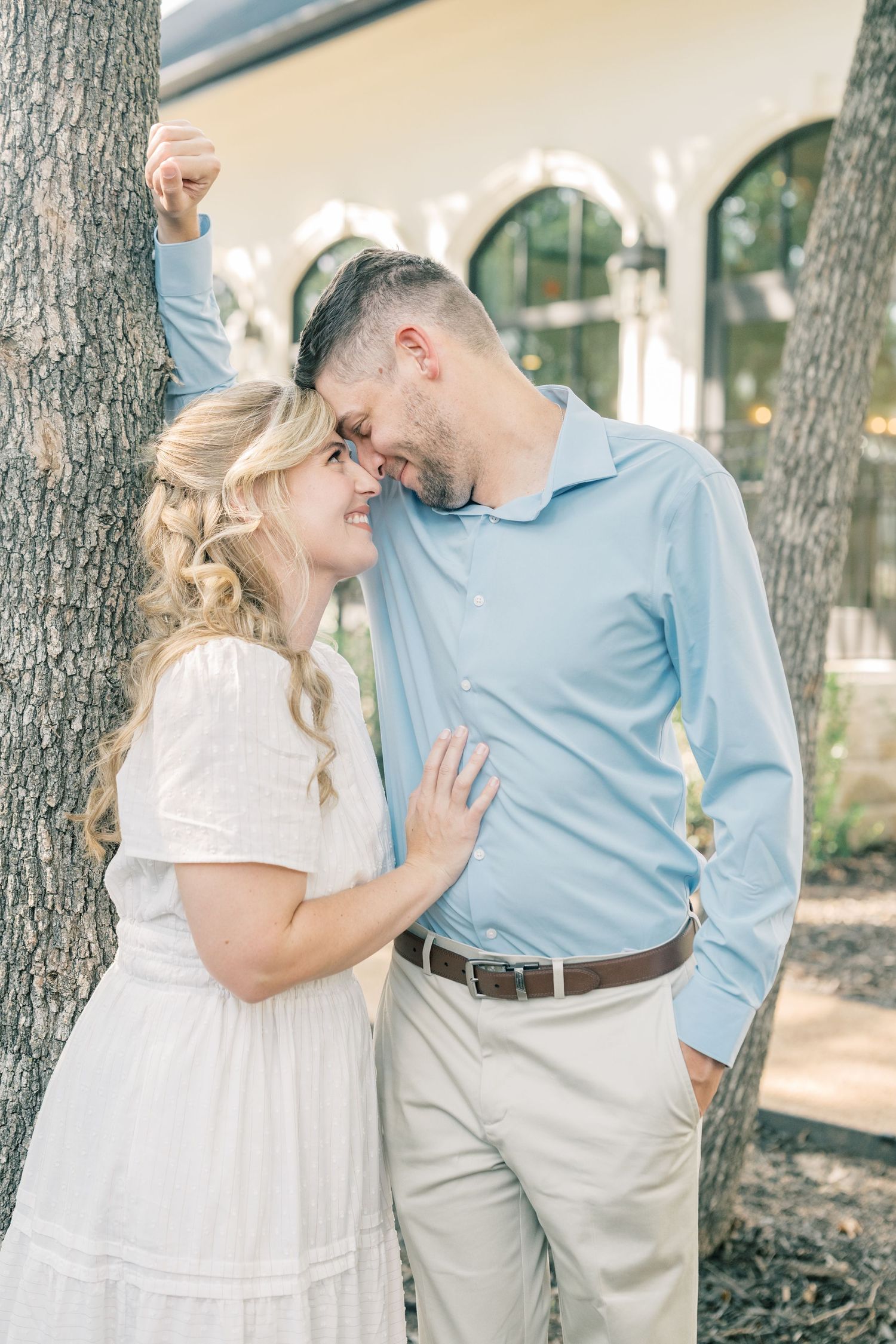 Smiling couple leaning foreheads together between two trees outside a white building with arched windows.
