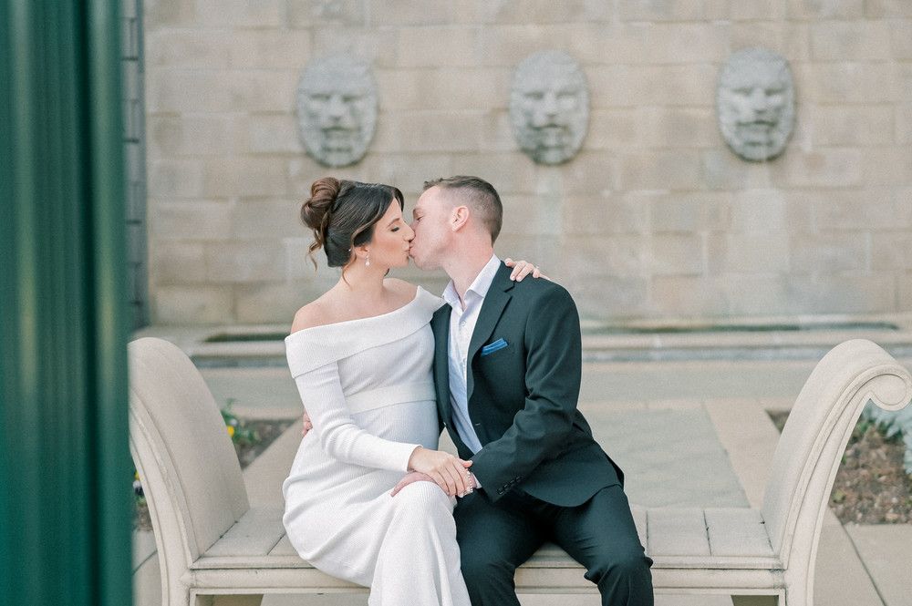 Bride and groom sharing a kiss while seated on stone bench before decorative wall with sculpted faces.