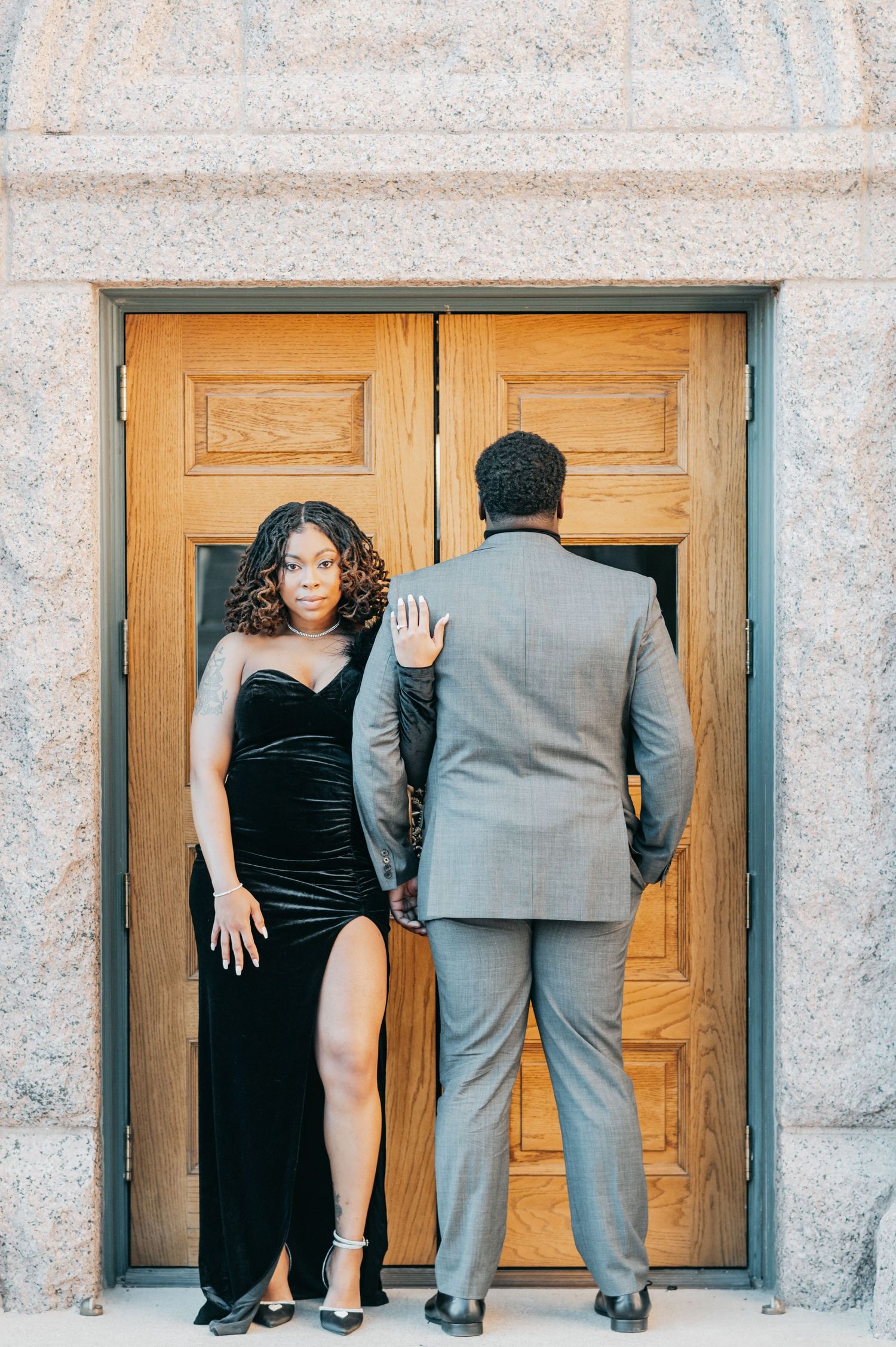 Woman in black velvet gown poses with man in gray suit at ornate wooden doors.