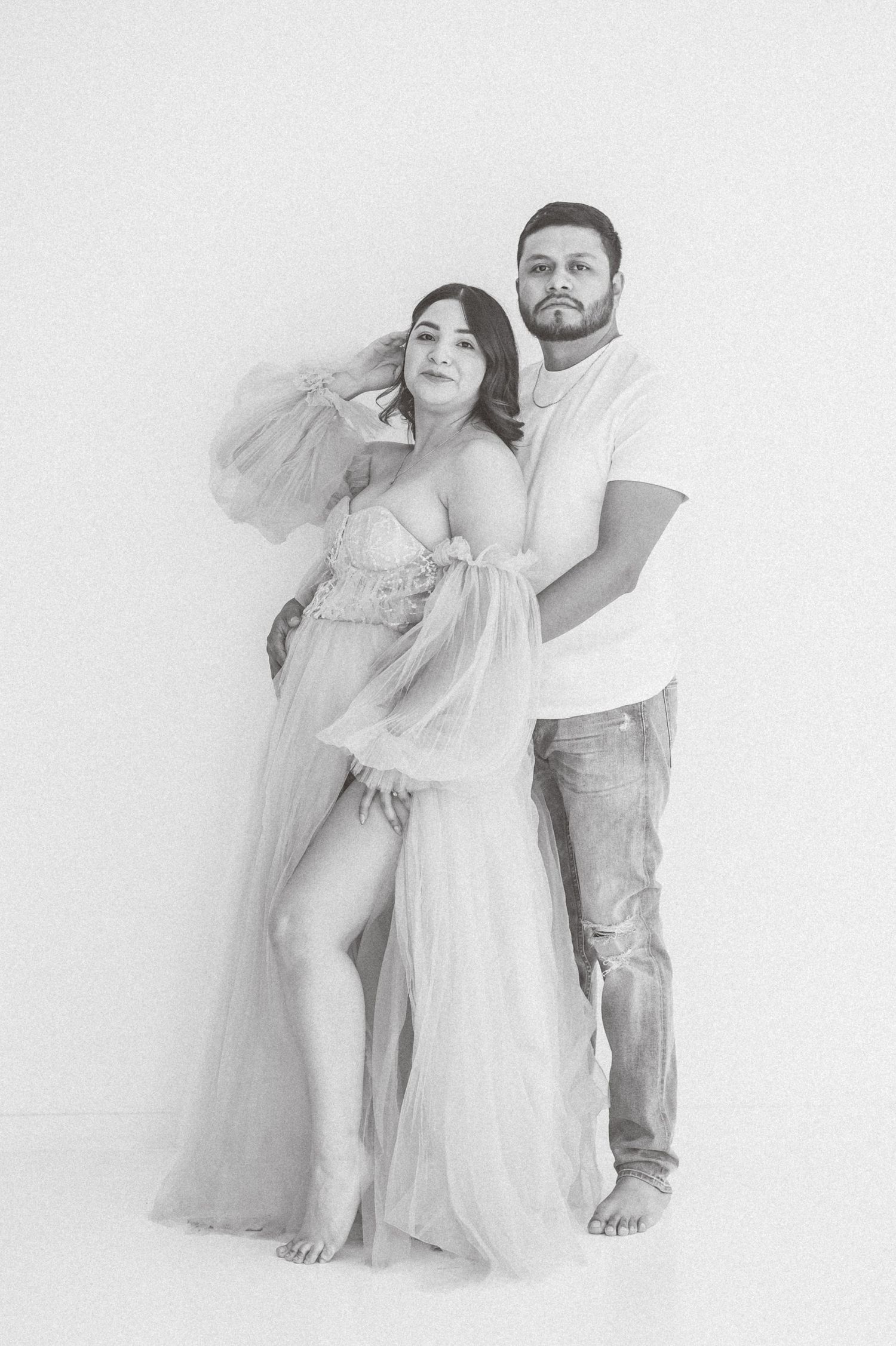 Black and white maternity photo of a couple, woman in sheer tulle gown, man in white tee and jeans, studio setting.