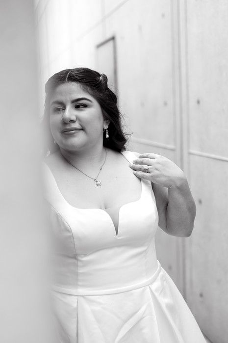 Black and white portrait of a bride in a deep V-neck wedding gown, leaning against a wall, smiling softly.