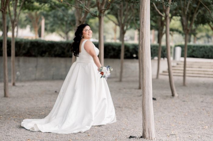 Bride in white ball gown holding bouquet poses among trees in outdoor garden setting.