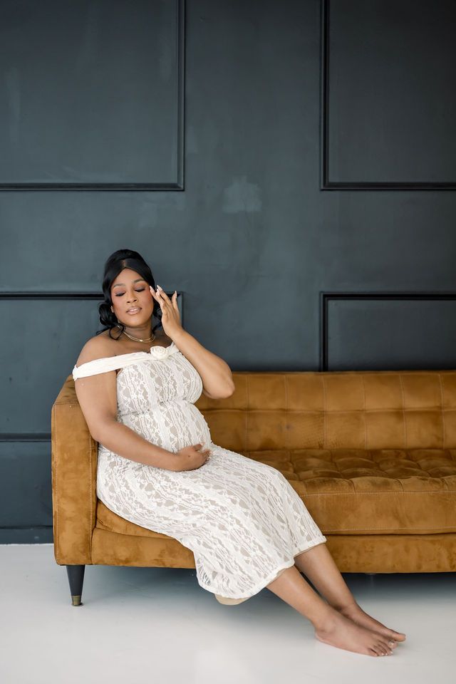 Pregnant woman in white lace off-shoulder dress sitting on camel sofa, touching face gracefully against dark wall.