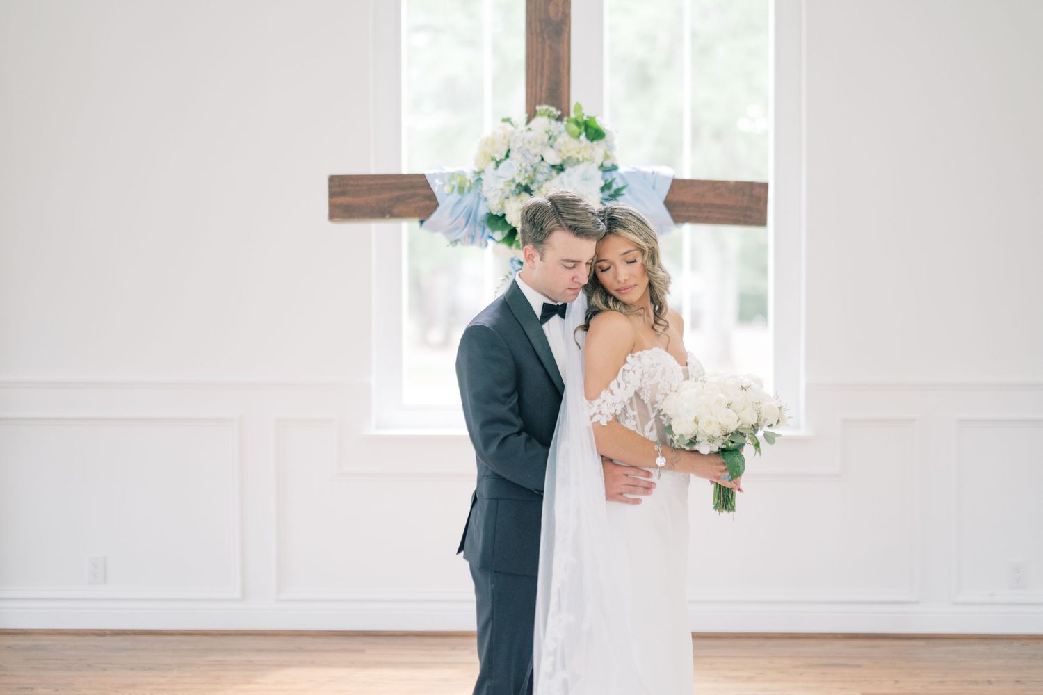 Bride and groom embracing tenderly in front of wooden cross adorned with white and blue flowers in chapel.