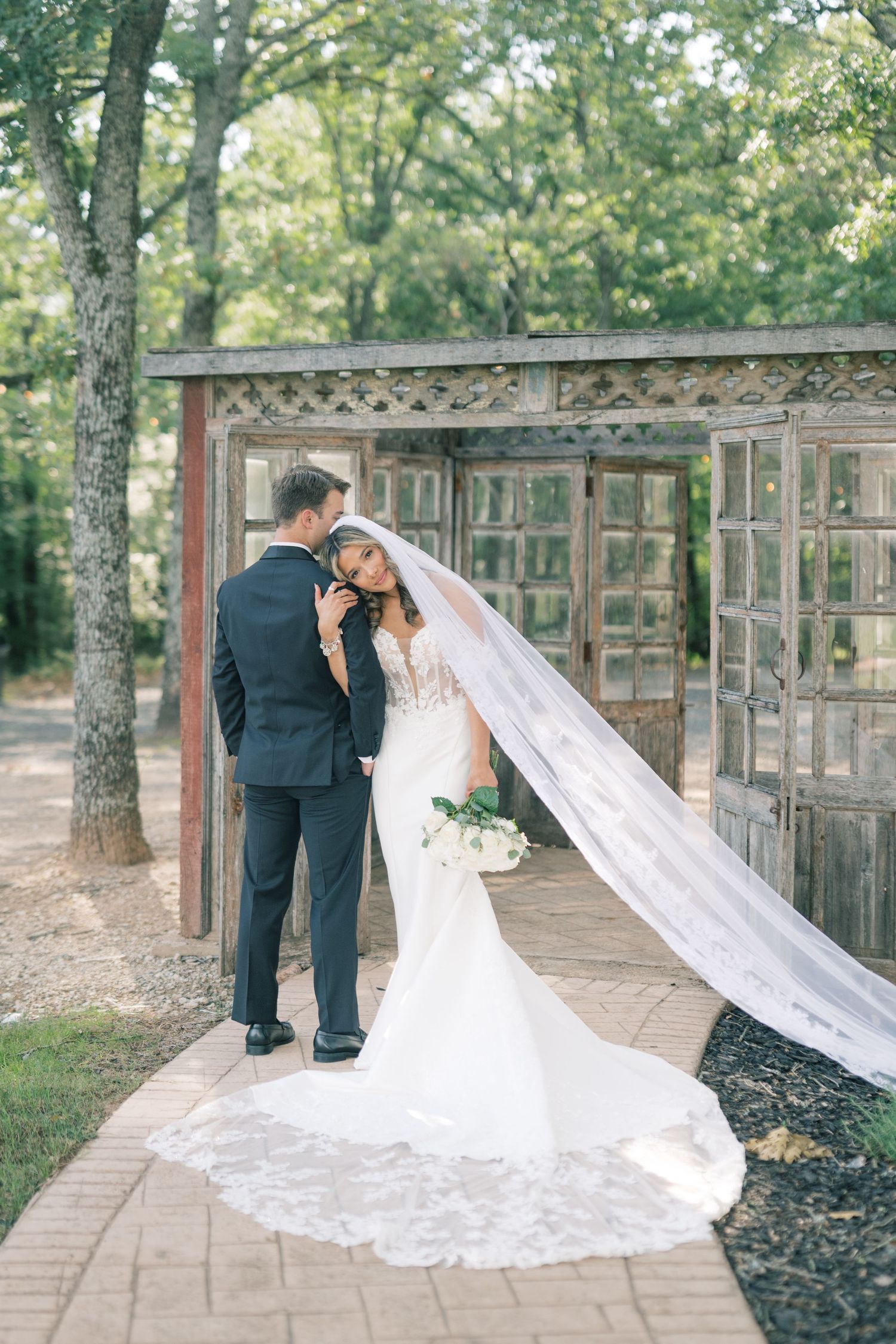 Bride and groom sharing a romantic moment near rustic wooden doors with long lace veil trailing behind.