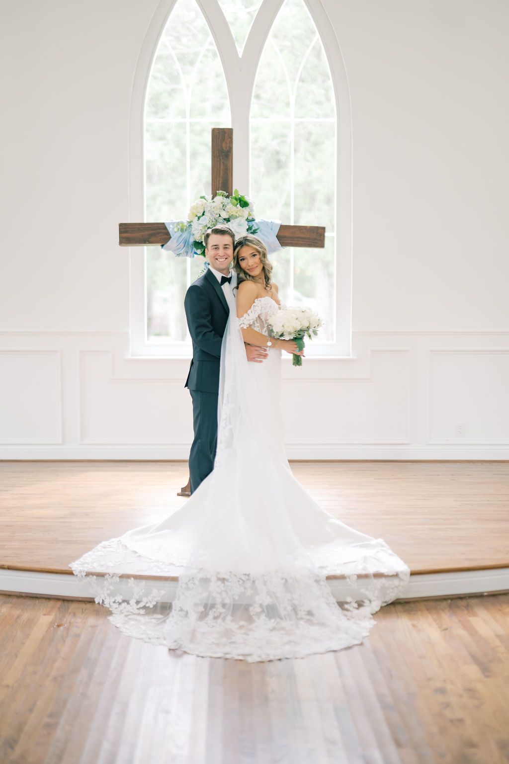 Bride and groom pose before a wooden cross in a bright white chapel, her lace train spread elegantly on hardwood floors.