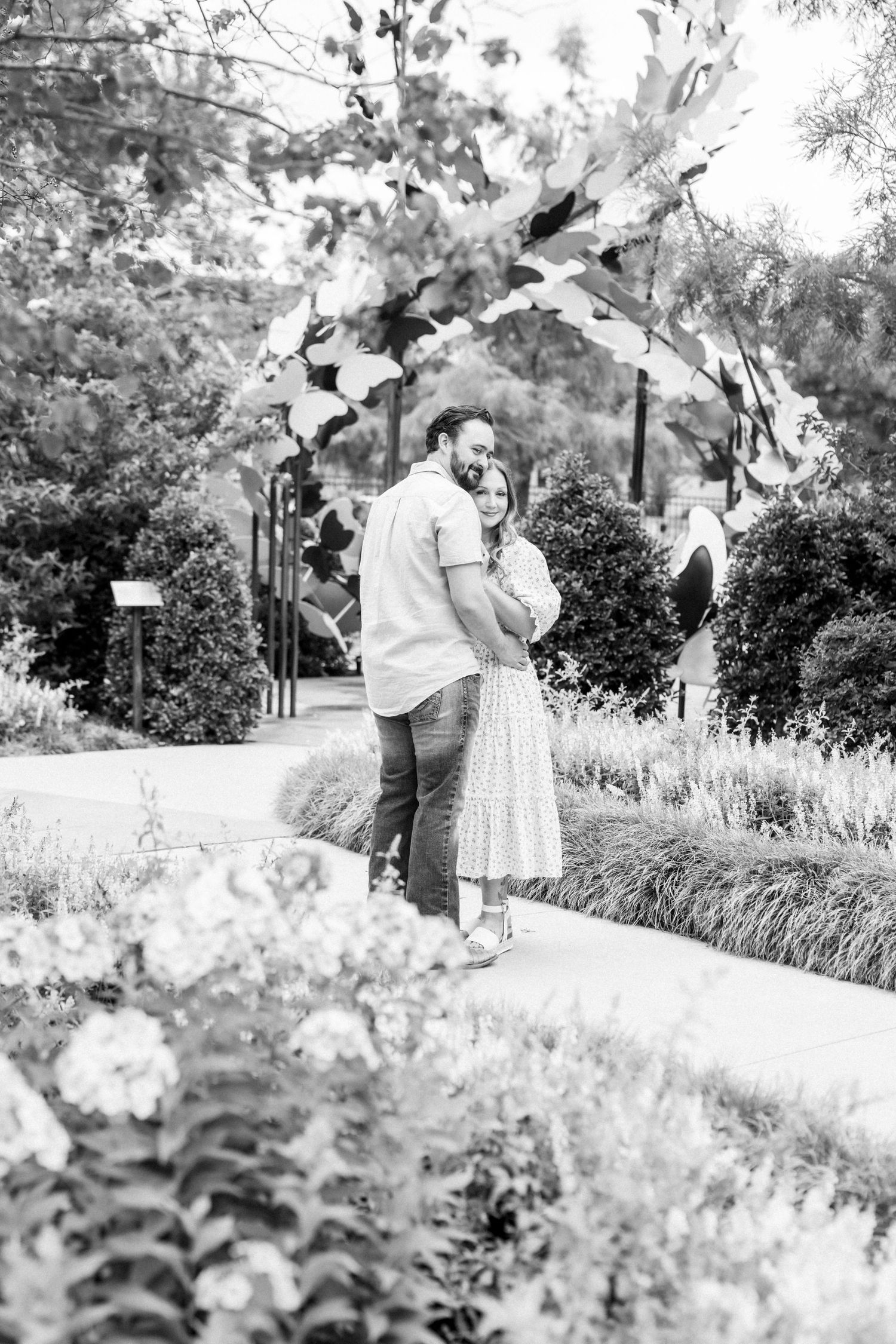 Black and white photo of a couple embracing in a lush garden pathway beneath a decorative floral arch.