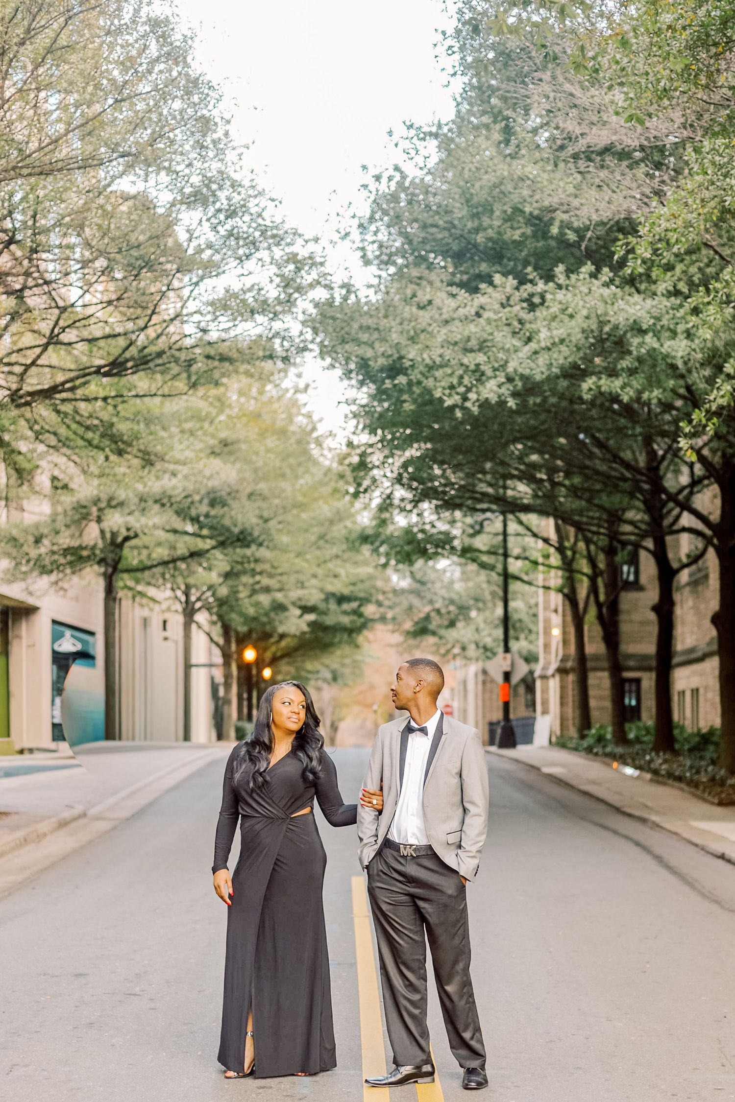 Elegant couple in formal attire standing on a tree-lined urban street, woman in black gown, man in gray suit.