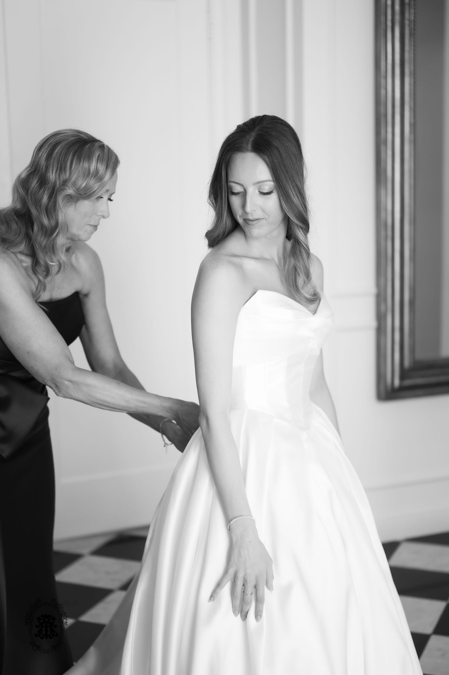 Black and white photo of a mother helping her daughter into a strapless white wedding gown before the ceremony.