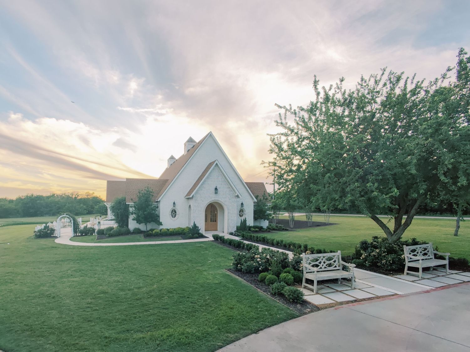 White chapel with arched entrance surrounded by lush green lawn and blooming trees at sunset.