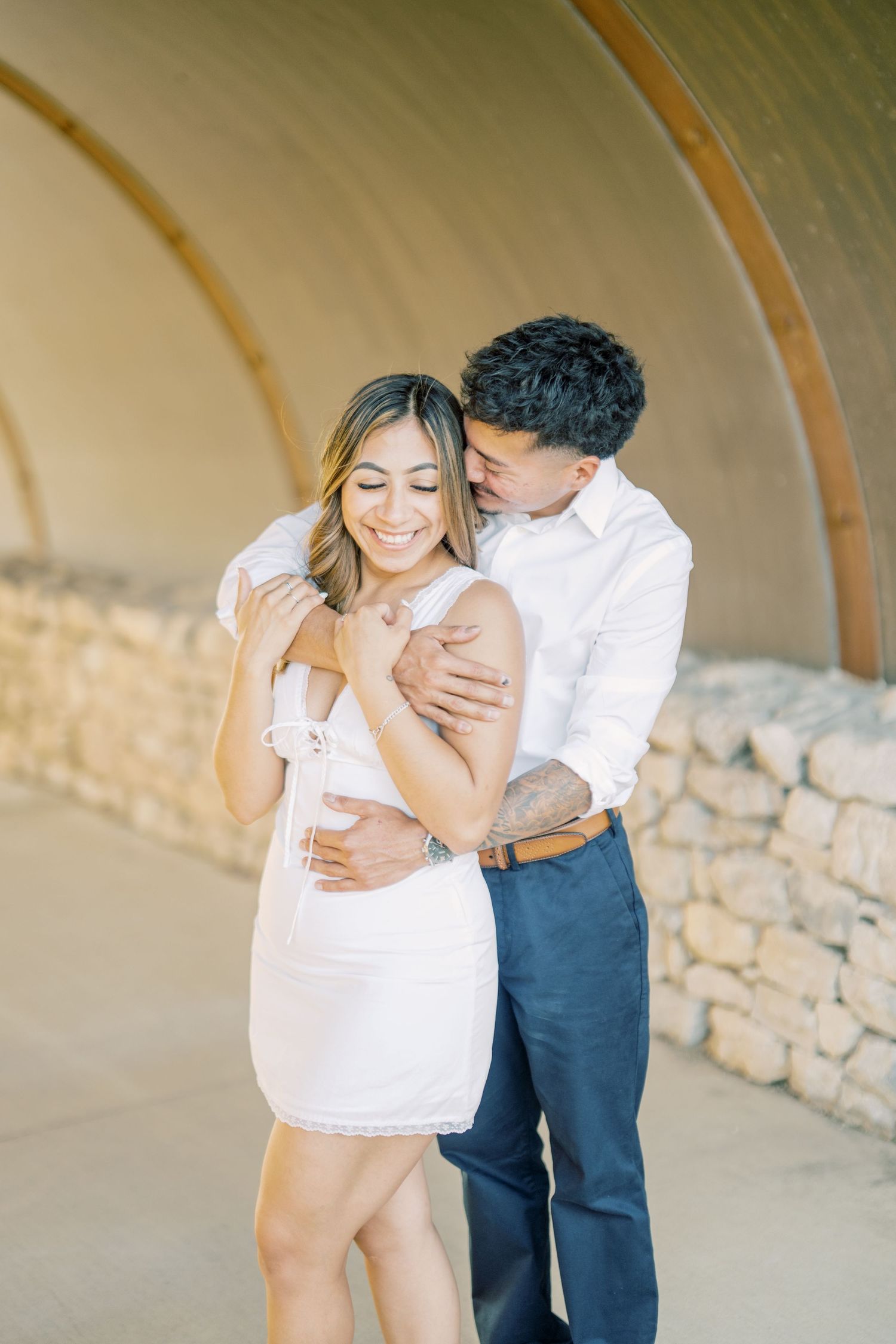 Couple sharing romantic embrace under stone archway, woman in white mini dress laughing joyfully during engagement photos.