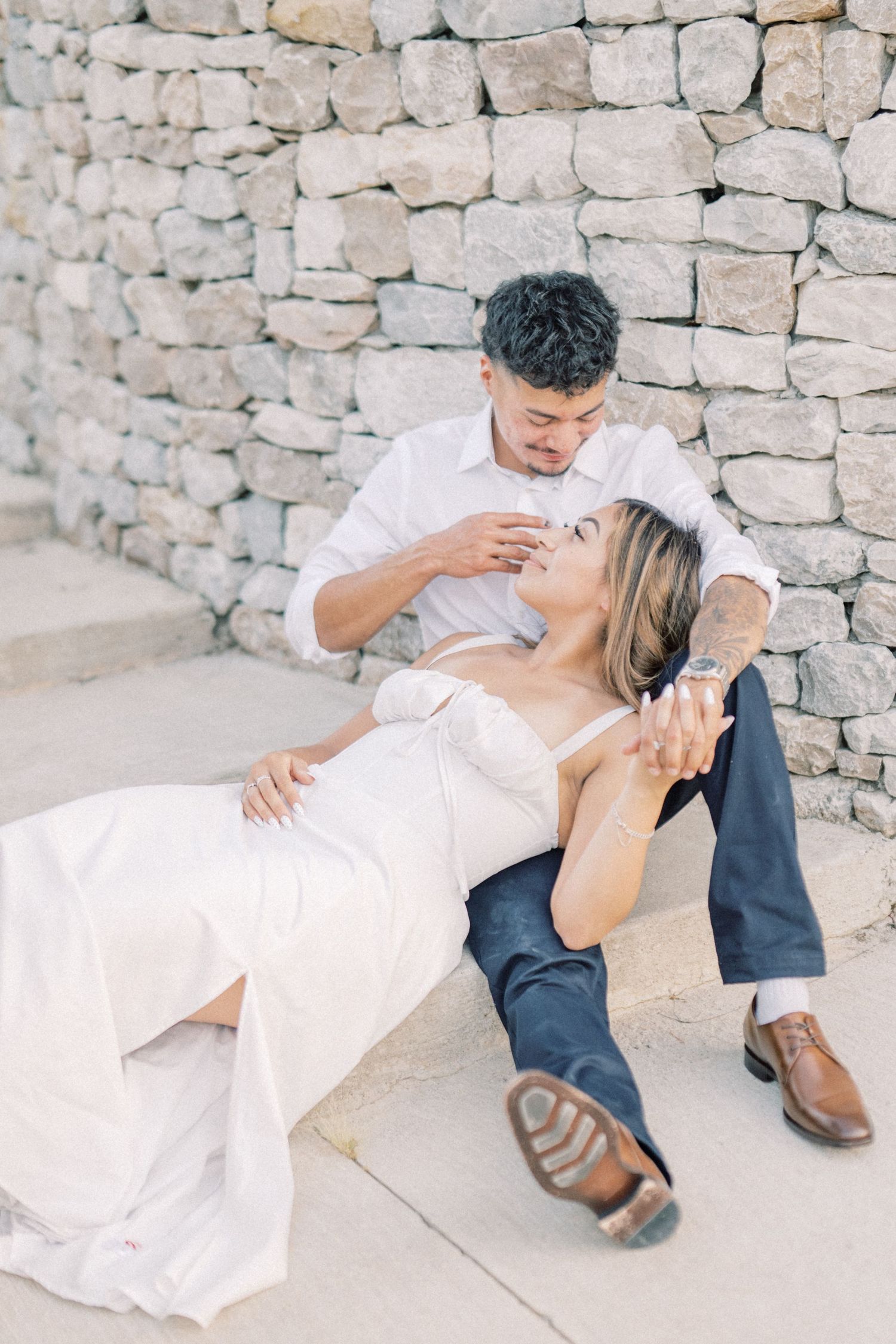 Couple sitting together against a stone wall, woman in white gown reclining against man in white shirt.