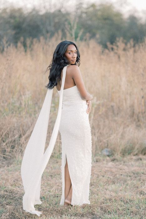 Elegant maternity portrait of woman in white dress with flowing fabric against rustic outdoor backdrop.