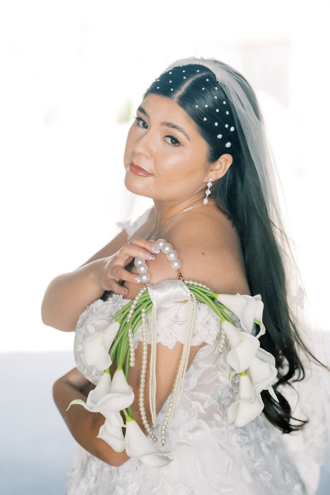 Bride in white dress adorned with pearls and white calla lilies poses elegantly against light background.